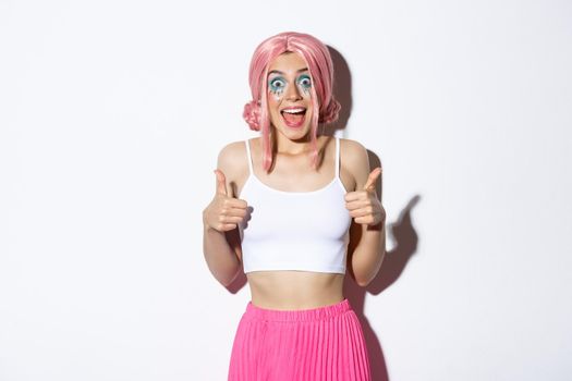 Portrait of satisfied smiling girl showing thumbs-up in approval, like something good, standing in halloween costume with pink with and bright makeup.