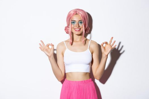 Portrait of smiling beautiful girl with pink anime wig and bright makeup, showing okay signs with satisfied expression, recommend something for halloween, standing over white background.