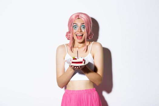 Portrait of excited birthday girl in pink wig, looking amused while making wish on b-day cake with lit candle, celebrating over white background.