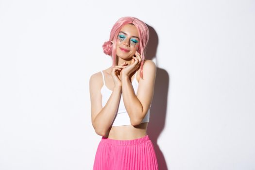 Portrait of tender stylish young girl in pink wig, close eyes and gently touching her face, daydreaming about something, standing over white background.