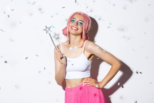 Image of beautiful happy girl in pink wig, celebrating halloween in fairy costume, holding magic wand and smiling while confetti floating in air.