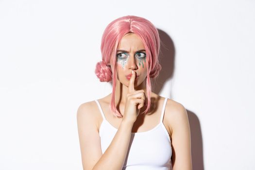 Close-up of worried attractive girl in pink anime hair, showing shush sign, press index finger to lips and looking concerned. Woman share a secret, standing over white background.