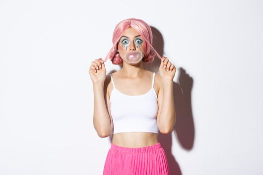 Portrait of attractive girl blowing pink bubble gum on face and looking silly, standing in anime wig and party outfit.