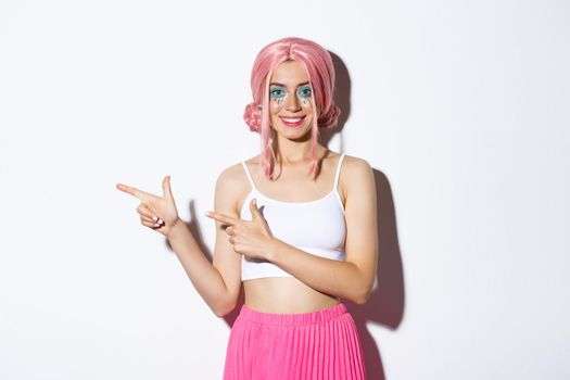 Image of trendy smiling girl dressed for halloween party, pointing fingers left, showing advertisement, standing over white background.