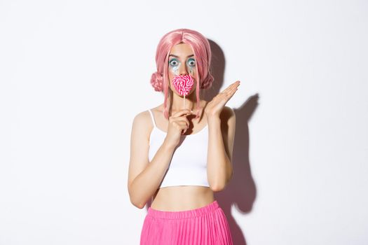 Image of flirty female model with pink wig and bright makeup, kissing heart-shaped candy and looking surprised, celebrating halloween.