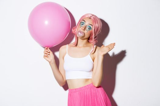 Image of pretty silly girl in pink wig, waving hand to say hello, holding balloon at party celebration, blowing bubble gum, standing over white background.