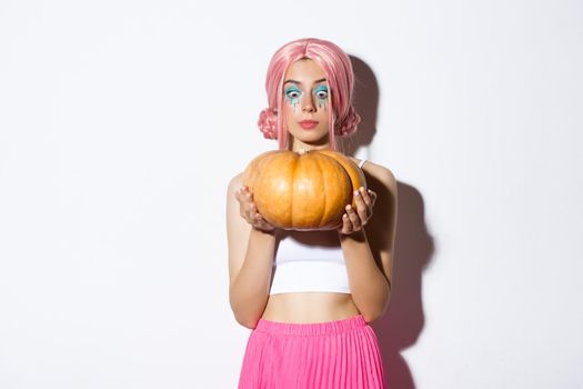 Image of pretty girl in pink wig and halloween makeup, looking excited at big pumpkin, standing over white background.