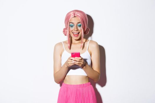 Portrait of surprised girl looking at happy news on mobile phone, wearing pink wig for party, halloween celebration, standing over white background.