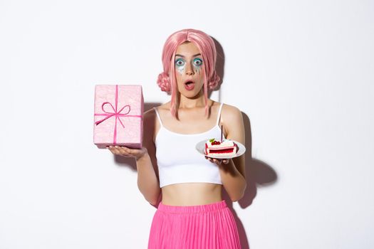 Portrait of surprised beautiful girl in pink wig, receive birthday gift, holding b-day cake and smiling happy, standing over white background.