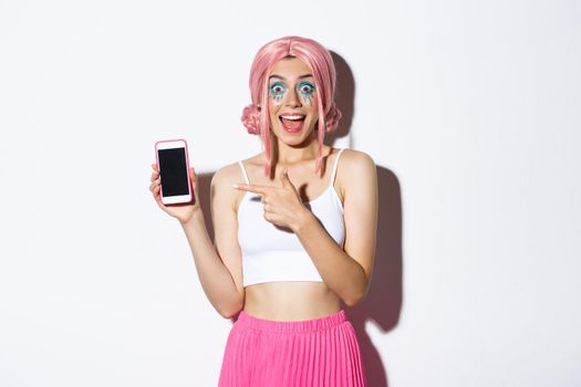 Portrait of excited beautiful girl in halloween costume, pink wig and bright makeup, pointing finger at mobile phone with amazed face, standing over white background .