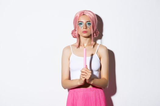 Image of serious looking girl dressed-up as fairy on halloween, holding candle, standing over white background in pink wig.