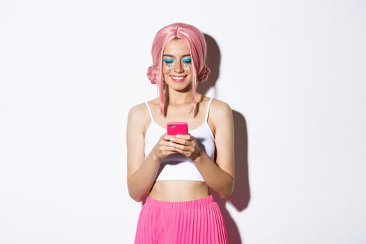Portrait of beautiful female with pink anime wig and bright makeup, dressed for party, smiling and looking at mobile phone, standing over white background.