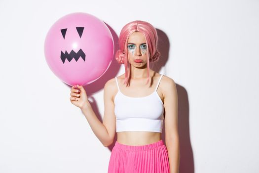 Image of sulking disappointed party girl with bright makeup, wearing pink wig, holding balloon with Jack-o'-lantern face, celebrating halloween.