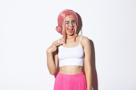 Image of cheeky pretty girl in pink wig and party outfit, winking and pointing at herself, wearing halloween costume, standing over white background.