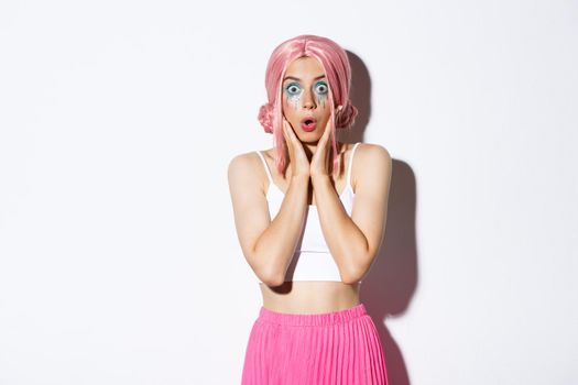 Portrait of surprised caucasian girl in pink wig and bright makeup, gasping amazed and stare at camera in awe, standing over white background in halloween party costume.