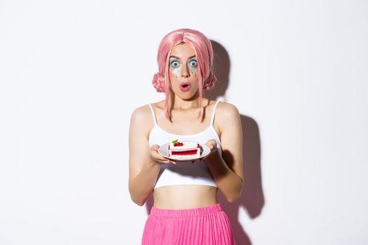 Image of surprised young birthday girl in pink wig, holding cake on plate and looking amazed at camera, standing over white background.