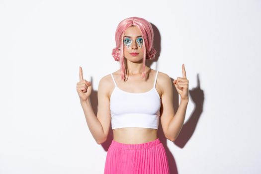 Portrait of stylish party girl with pink anime wig and halloween makeup, pointing fingers up at your logo, standing over white background.