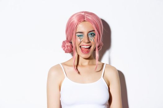 Close-up of pretty coquettish girl in pink wig, winking at camera and smiling, standing over white background.