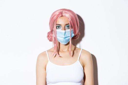 Close-up of girl in pink wig and medical mask, celebrating halloween during coronavirus pandemic, standing over white background.
