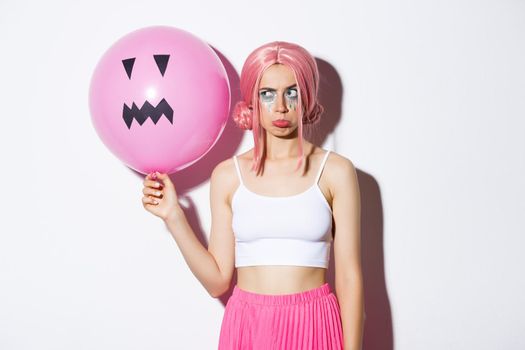 Moody cute girl in pink wig, pouting jealous or displeased, standing with jack-o-lantern balloon, celebrating halloween, standing over white background.