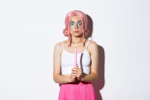 Image of scared girl in pink wig looking shocked at camera, standing with lit candle over white background, celebrating halloween.