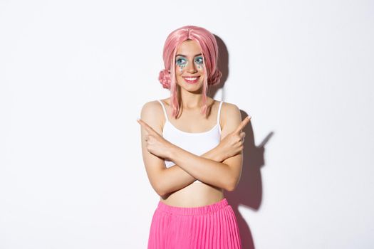 Beautiful smiling party girl in halloween costume and pink wig, pointing fingers sideways but looking left, making choice, standing over white background.