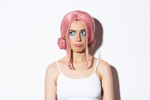 Close-up of confused cute girl in pink wig, grimacing and looking upper left corner with disappointed face, standing in halloween costume over white background.