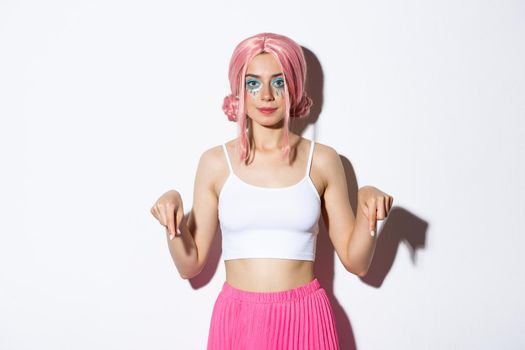Serious and confident caucasian woman in pink anime wig and party makeup, pointing fingers down, showing advertisement about holiday celebration.