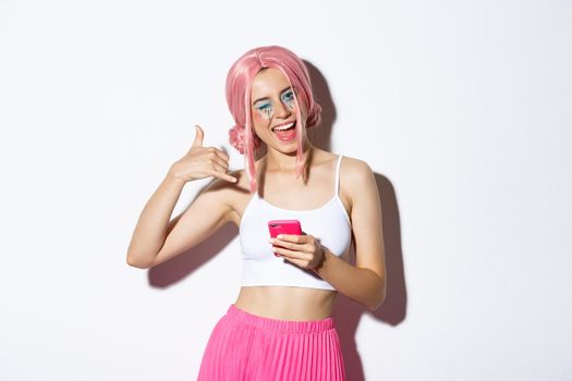 Cheerful party girl making phone call sign and winking flirty at camera, holding smartphone, standing in pink wig over white background.