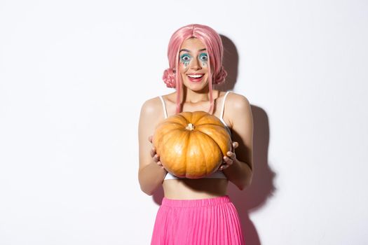 Portrait of happy girl looking excited for halloween party, holding pumpkin, wearing cute pink wig, standing over white background.