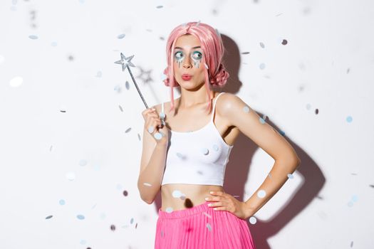 Image of beautiful happy girl in pink wig, celebrating halloween in fairy costume, holding magic wand and smiling while confetti floating in air.