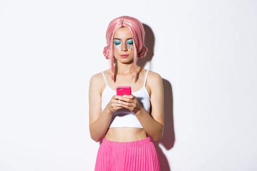 Image of stylish glamour girl in pink wig, looking serious at mobile phone, standing in party outfit over white background.