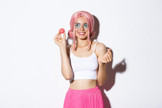 Portrait of lovely female model eating macaroons and smiling, wearing pink wig and outfit for party, standing over white background.