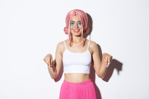 Beautiful smiling party girl with pink hair and bright makeup, pointing fingers down at your logo, wearing halloween costume, standing over white background.