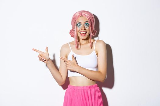 Portrait of excited happy girl in pink wig and bright makeup celebrating something, pointing fingers left at your logo about holidays and parties, standing over white background.