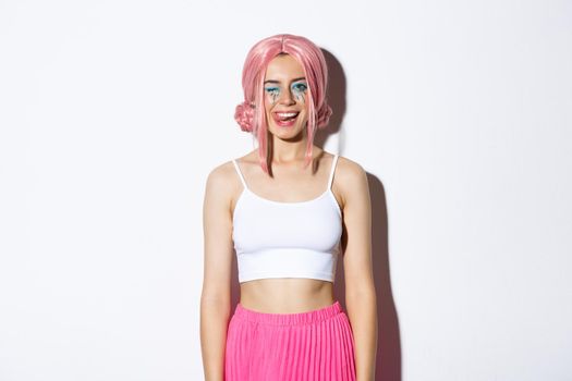 Portrait of carefree smiling female model in pink party wig, winking and showing tongue, celebrating halloween, standing over white background.