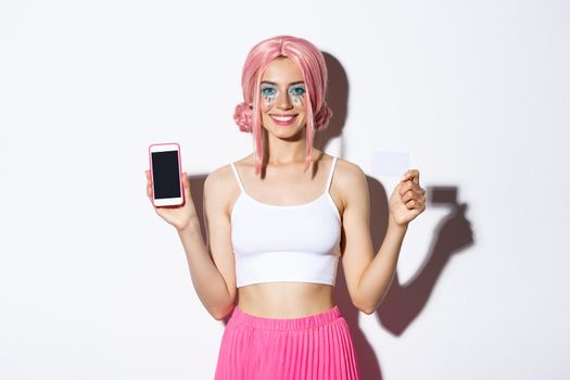 Image of smiling beautiful girl in pink wig, with bright makeup, showing credit card and mobile phone screen, standing over white background.