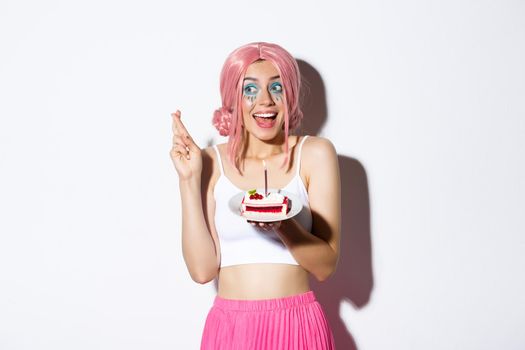 Portrait of hopeful birthday girl in pink wig, making wish with fingers crossed, holding b-day cake, standing over white background.