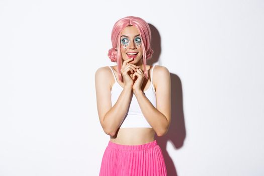 Portrait of intrigued beautiful girl with pink wig and bright makeup, looking left at something tempting, standing in halloween costume over white background.