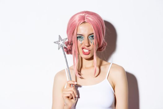 Close-up of sassy attractive woman in pink wig, holding magic wand, cosplay fairy at halloween party, standing over white background.