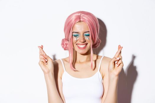 Close-up of excited girl with pink wig, making wish with fingers crossed, smiling hopeful, wearing halloween costume.