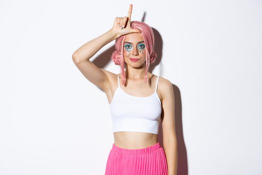 Image of arrogant young stylish woman with pink hair and halloween makeup, showing loser sign on forehead as mocking someone, standing over white background.