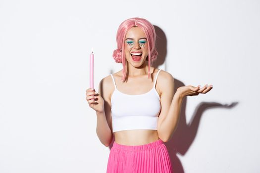 Portrait of happy cute girl with pink candle, wearing halloween costume and laughing, standing over white background.