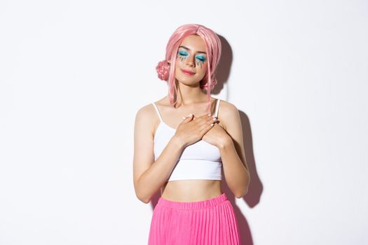 Portrait of nostalgic beautiful young woman in pink wig, bright makeup, close eyes and holding hands on heart, remember something or daydreaming, standing over white background.