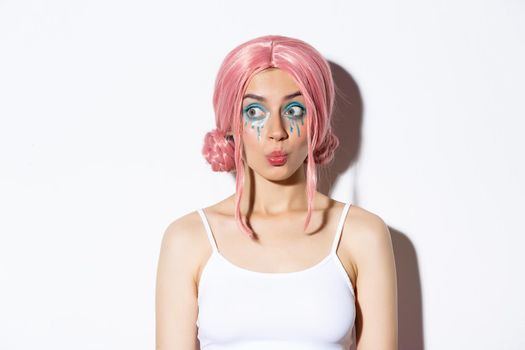 Image of beautiful silly girl in pink wig, with bright makeup, pouting and looking interested left, standing over white background.