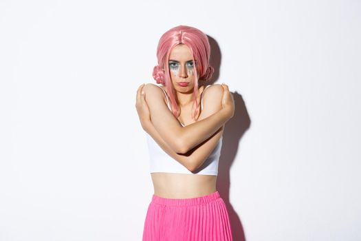 Portrait of offended cute girl hugging herself and sulking, feeling insulted or sad about something, wearing pink wig for halloween celebration, standing over white background.