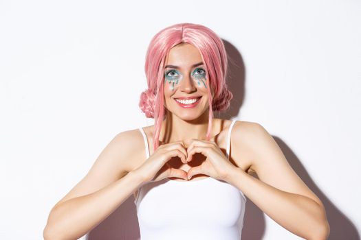 Close-up of hopeful smiling girl in pink party wig, looking up and showing heart sign, standing over white background.