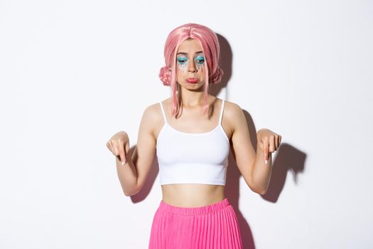 Portrait of gloomy cute girl in pink wig and party outfit, looking disappointed and pouting, pointing fingers down at bad news, standing over white background.