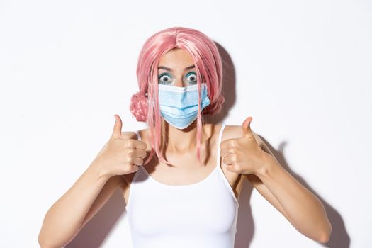 Close-up of excited cute girl in pink wig and medical mask, showing thumbs-up with happy expression, celebrating halloween during covid-19 pandemic.