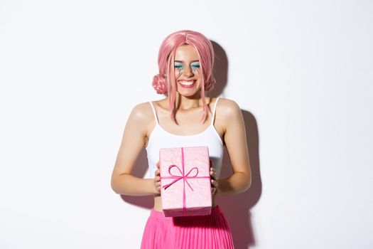 Portrait of happy beautiful b-day girl looking excited, receiving birthday gift and smiling joyfully, standing in pink wig and party outfit over white background.